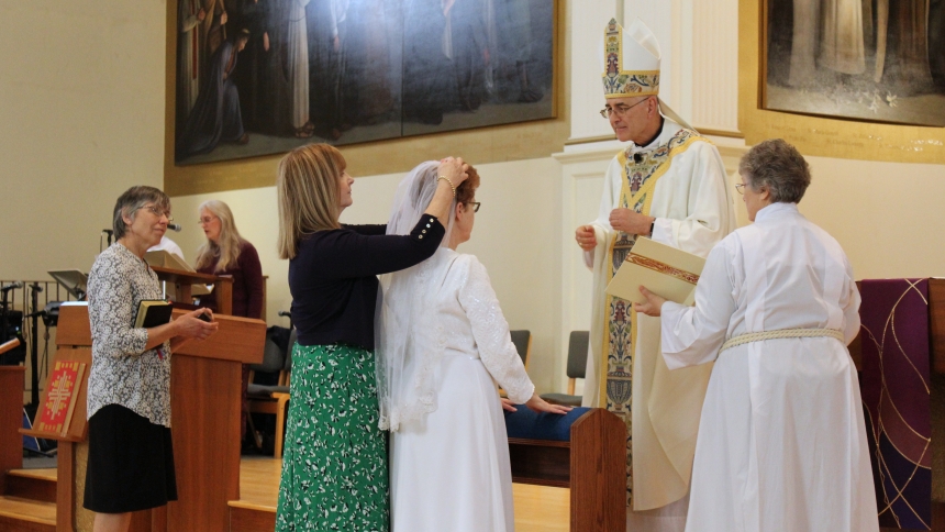 woman putting veil on another woman's head during religious ceremony with a bishop