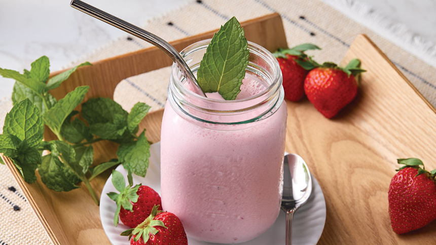 A strawberry milkshake in a mason jar atop a wooden tray, garnished with whole strawberries and mint.